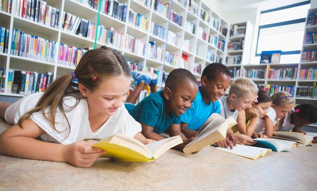 crianças de diferentes etnias deitadas no chão de uma biblioteca escolar, lendo livros com entusiasmo.