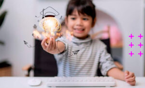 garotinha sorridente sentada em frente a uma mesa com teclado e mouse segurando uma lâmpada brilhando em uma das mãos.