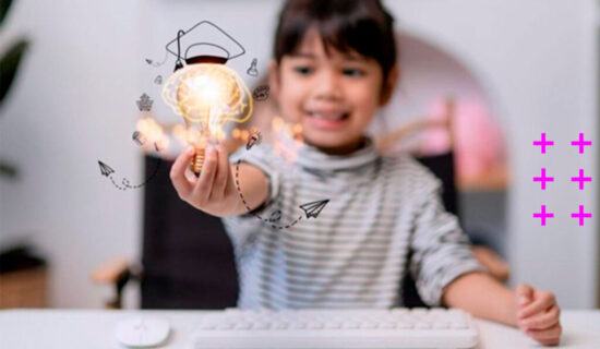 garotinha sorridente sentada em frente a uma mesa com teclado e mouse segurando uma lâmpada brilhando em uma das mãos.