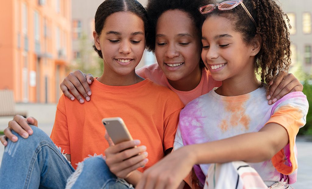 Mãe abraçando suas duas filhas enquanto as meninas mostram para ela algo no celular.