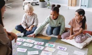 Professora sentada no chão com alunos do ensino fundamental aplicando atividade educativa em inglês.