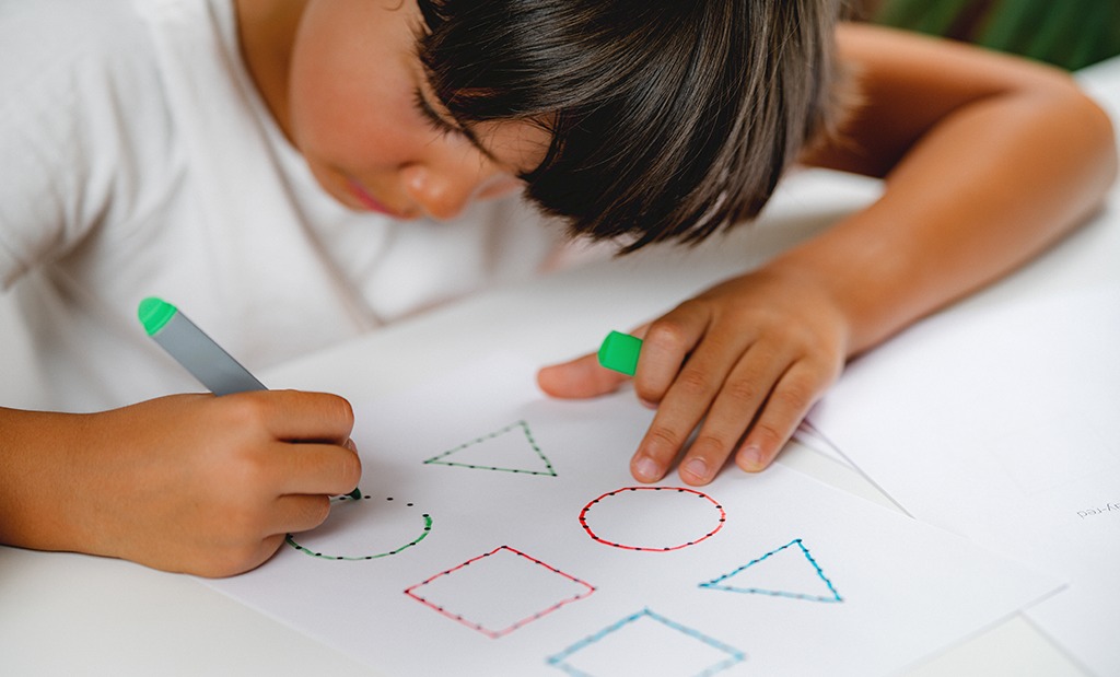 Aluno do ensino fundamental completando desenhos pontilhados com caneta que formam formas geométricas.