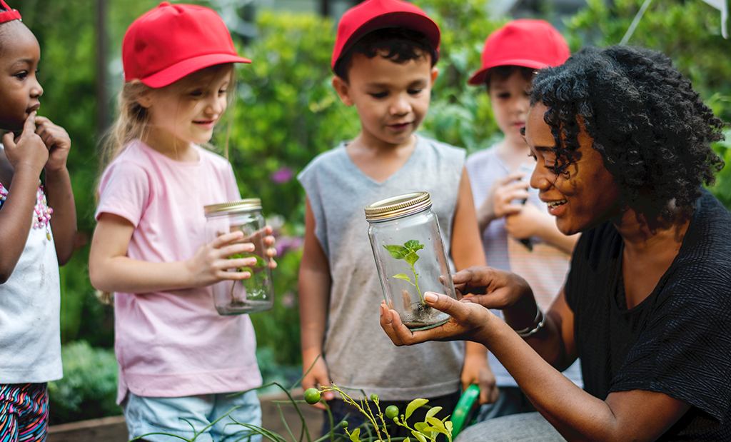 Professora e alunos em um parque com área verde olhando para potes de vidro com plantas.