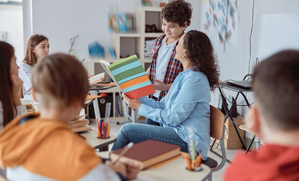 Professora no centro de uma roda de alunos aplicando a metodologia de sala de aula invertida.