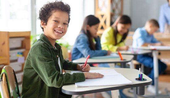 Menino em sala de aula escrevendo em um caderno e sorrindo para representar o ensino fundamental.