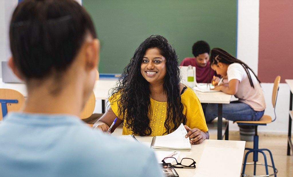 Aluna do ensino médio em sala de aula conversando com colega de classe enquanto faz anotações no caderno.