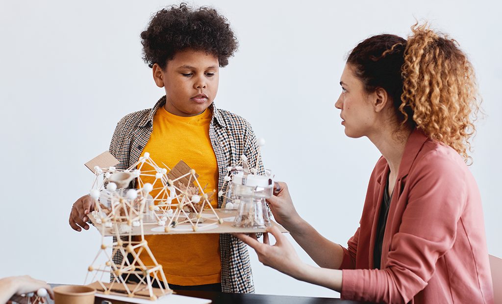 Professora aplicando cultura Maker em sala de aula com aluno que está criando uma maquete de ciências.