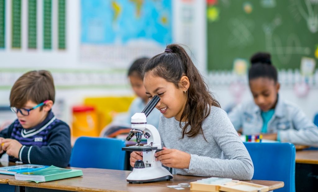 Crianças em uma sala de aula participando de uma atividade científica. Em destaque, uma menina sorrindo enquanto olha através de um microscópio, com outras crianças ao fundo concentradas em suas atividades.