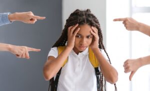 Uma menina com tranças e mochila amarela parece angustiada, segurando a cabeça com as mãos. Vários dedos apontam para ela, sugerindo um ambiente de pressão ou crítica.