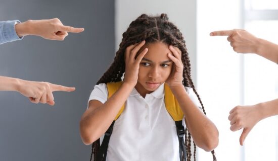 Uma menina com tranças e mochila amarela parece angustiada, segurando a cabeça com as mãos. Vários dedos apontam para ela, sugerindo um ambiente de pressão ou crítica.