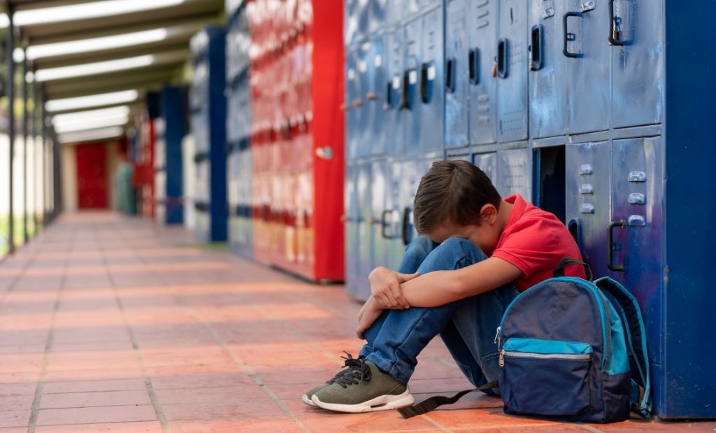 Uma menino sentado no chão de uma ambiente escolar, ao lado de sua mochila, apoiando a cabeça nos joelhos, parecendo estar triste e frustrado. Atrás dele há escaninhos azuis.