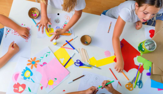 Crianças reunidas em volta de uma mesa, desenhando e colorindo em papéis com canetinhas coloridas.