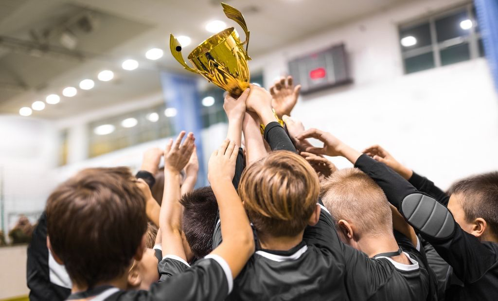 Um grupo de meninos com uniforme de futebol comemoram a vitória no jogo levantando um troféu.