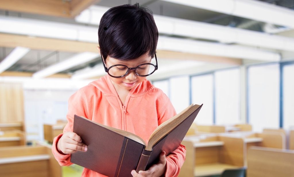 Menino de óculos com aproximadamente 8 anos, usando um moletom laranja e segurando um livro marrom, em uma sala de leitura.