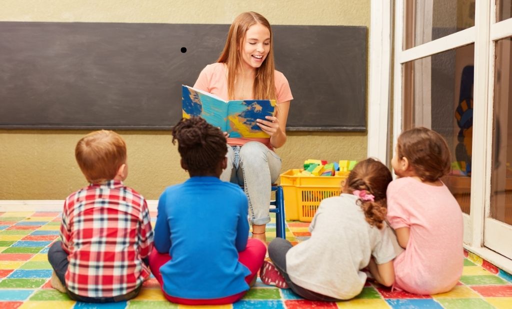 Professora com uma blusa rosa lendo um livro para quatro crianças sentadas em sua frente.