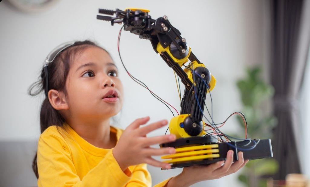 Menina segurando um braço robótico preto e amarelo, olhando atentamente para o dispositivo enquanto explora seu funcionamento.