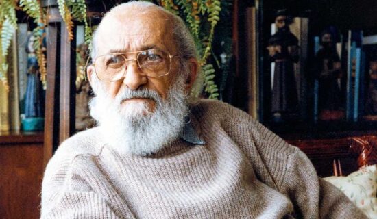 Homem calvo com barba branca e robusta, usando óculos de grau e um suéter bege. Ao fundo, plantas, livros e esculturas de santos.