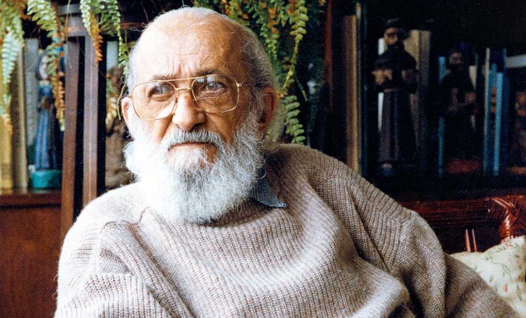 Homem calvo com barba branca e robusta, usando óculos de grau e um suéter bege. Ao fundo, plantas, livros e esculturas de santos.