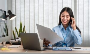 Mulher sorridente vestindo uma camisa azul e usando um crachá, sentada à mesa de trabalho. Ela está ao telefone e segurando um documento. Na mesa, há um laptop, uma luminária, alguns papéis e uma planta decorativa ao fundo.