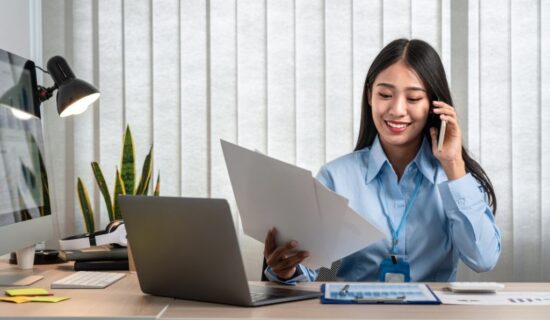 Mulher sorridente vestindo uma camisa azul e usando um crachá, sentada à mesa de trabalho. Ela está ao telefone e segurando um documento. Na mesa, há um laptop, uma luminária, alguns papéis e uma planta decorativa ao fundo.