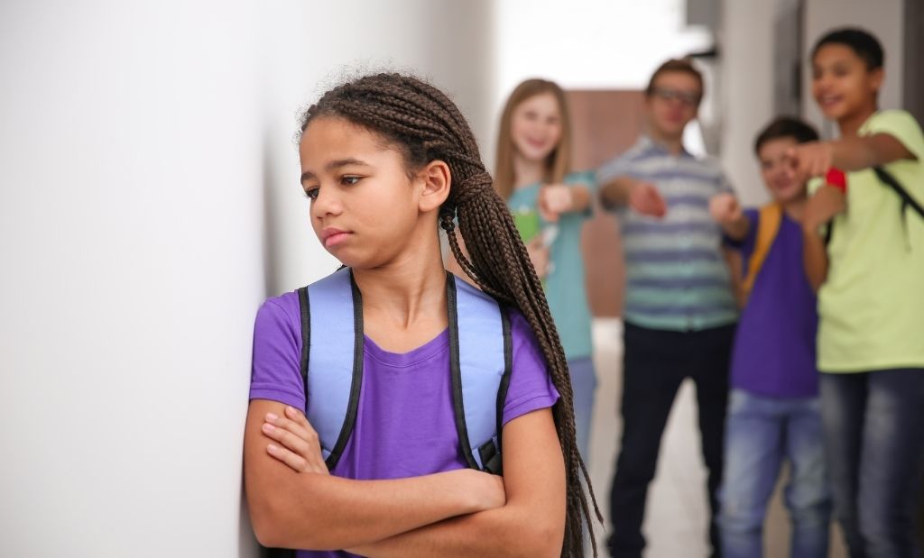 estudante isolada em um corredor, vítima de bullying por colegas ao fundo.